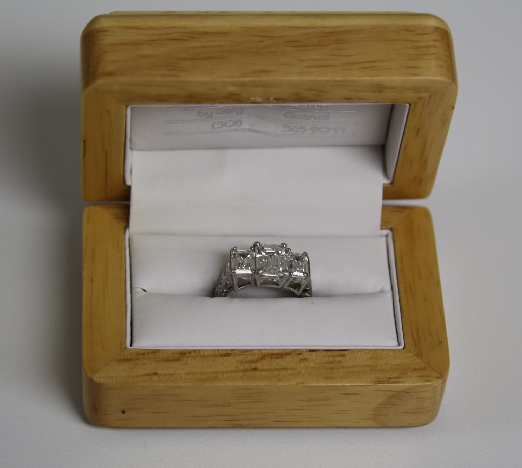 A silver ring with three square-cut stones displayed in an open light wood ring box.