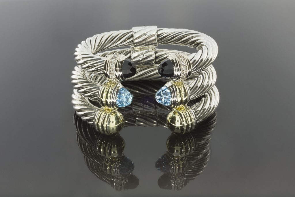 A close-up of an intertwined Yurman bracelet featuring blue and black gemstones, reflecting elegantly on a dark surface.