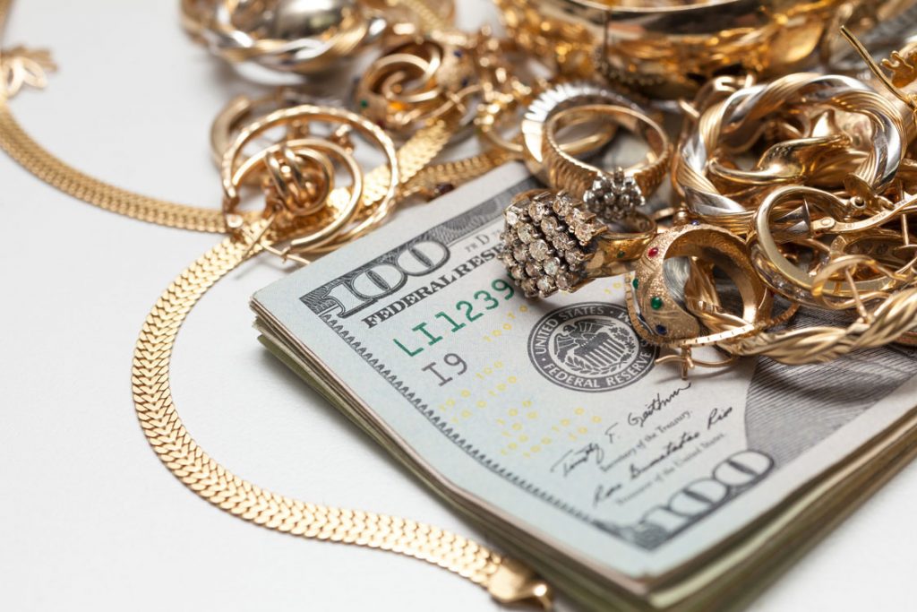 A stack of hundred-dollar bills surrounded by an assortment of gold jewelry, including rings, necklaces, and earrings.