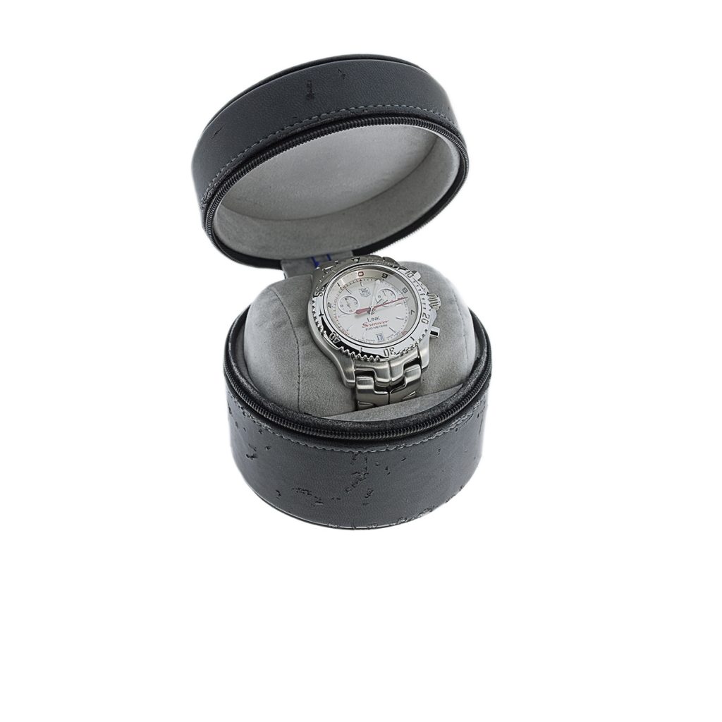 A silver watch with a white face rests inside a black, round leather case with a grey cushion. The case is partially open.