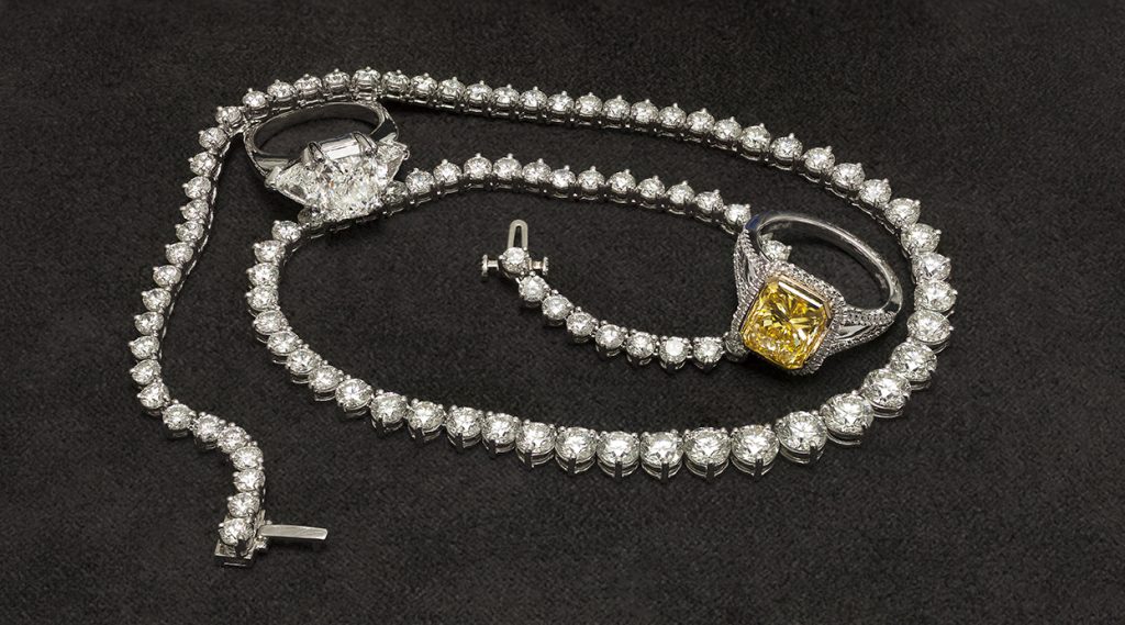 A full diamond necklace and two diamond rings, one with a yellow stone on a black surface.