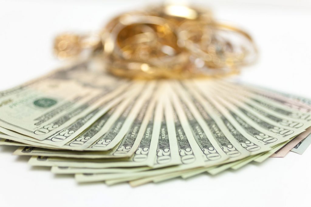 A fan of $20 bills with gold jewelry blurred in the background on a white surface.