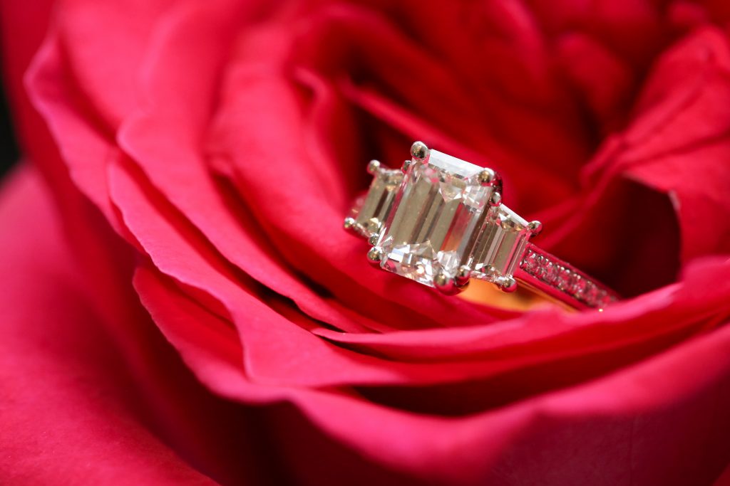 A stunning diamond engagement ring featuring three emerald-cut stones, elegantly resting on vibrant red rose petals.
