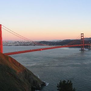 The Golden Gate Bridge spans across the water at sunset, with the city of San Francisco visible in the background and hills in the foreground