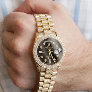 A close-up of a hand holding a luxurious gold Rolex Day-Date watch with a black dial, diamond accents, and a polished gold bracelet.