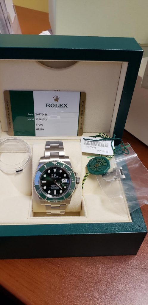 Close-up of a Rolex watch in a green box, with a warranty card and tags, showcasing a black dial and silver bracelet.