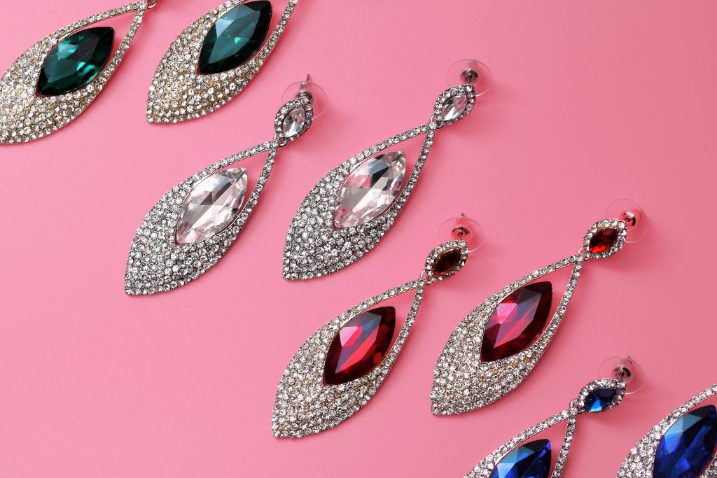 Four pairs of jeweled earrings with different colored gemstones—green, clear, red, and blue—are arranged in rows on a pink background.