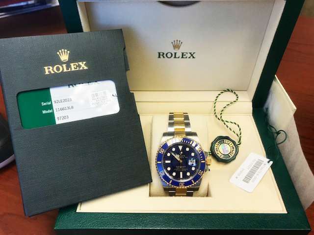 A Rolex watch with a blue dial and gold accents, displayed in its green box alongside a warranty card and price tag.
