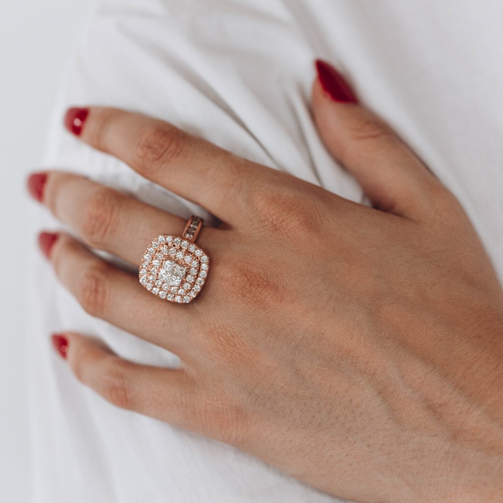 A woman in a white shirt is holding her arm, showing her hand with a red nail polish and wearing a rose gold engagement ring