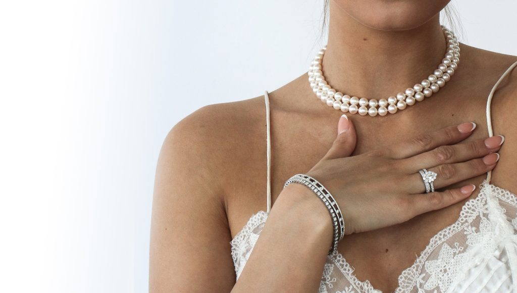 A close-up of a woman's neck and hands adorned with a pearl choker, a sparkling bracelet, and an elegant ring, against a white background.