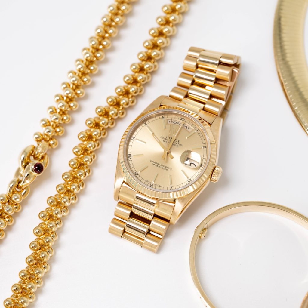 A luxurious gold Rolex watch next to a golden beaded necklace and a gold bangle on a white surface, exuding elegance and richness.