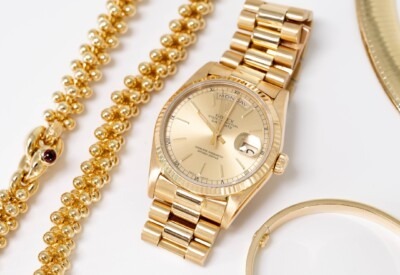 A luxurious gold Rolex watch next to a golden beaded necklace and a gold bangle on a white surface, exuding elegance and richness.