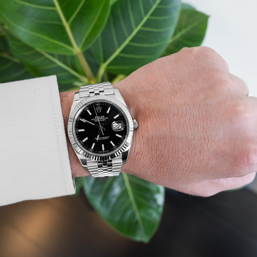 A person wearing a silver wristwatch with a black face, standing in front of a green leafy plant.