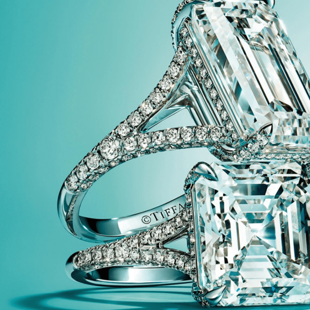A close-up view of exquisite Tiffany & Co. diamond engagement rings featuring radiant emerald-cut diamonds set on sparkling pavΓ© diamond bands, against the iconic Tiffany blue background.