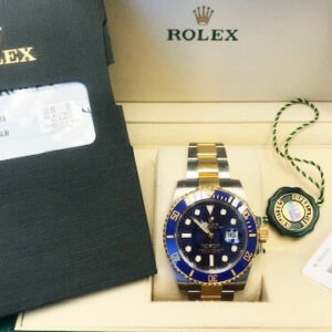 A Rolex watch with a blue dial and gold accents, displayed in its green box alongside a warranty card and price tag.