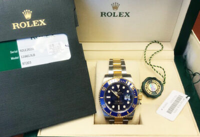 A Rolex watch with a blue dial and gold accents, displayed in its green box alongside a warranty card and price tag.