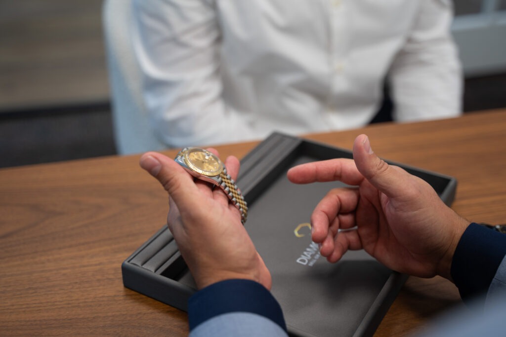 A person holds a gold wristwatch over a display tray on a wooden table, while another person in a white shirt sits across from them at Diamond Banc.
