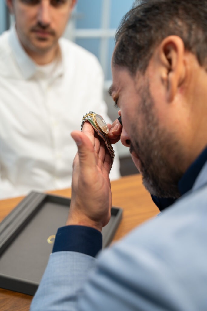 Diamond Banc staff examines a wristwatch closely with a magnifier while another man sits across the table.