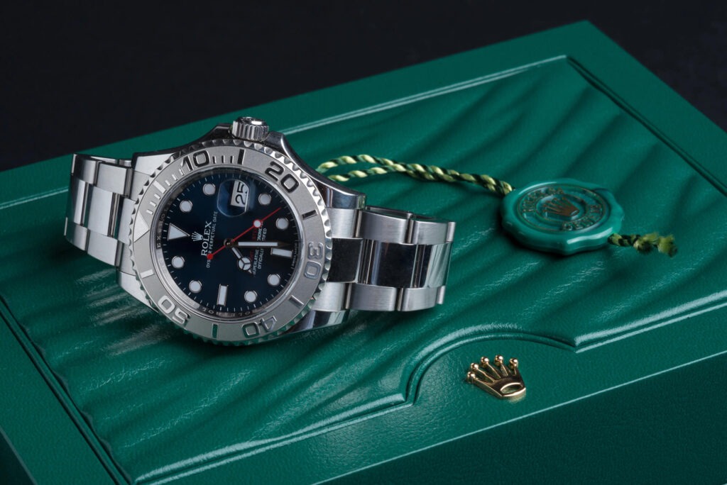 A silver Rolex Yacht-Master watch with a black dial rests on a green Rolex box, accompanied by a green seal and gold crown emblem.