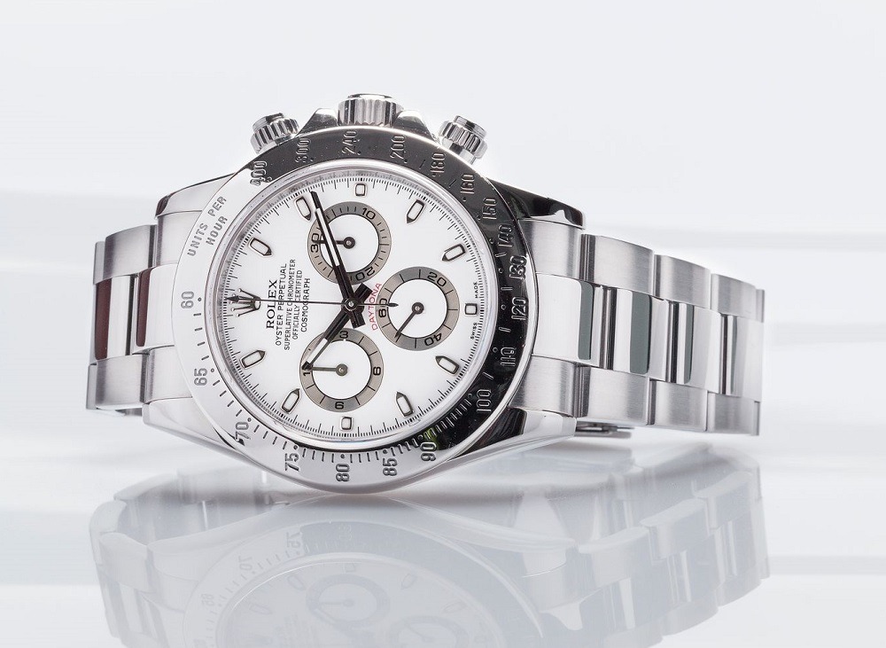 	
A stainless steel Rolex Daytona watch with a white dial and three subdials, lying on its side against a reflective white background.