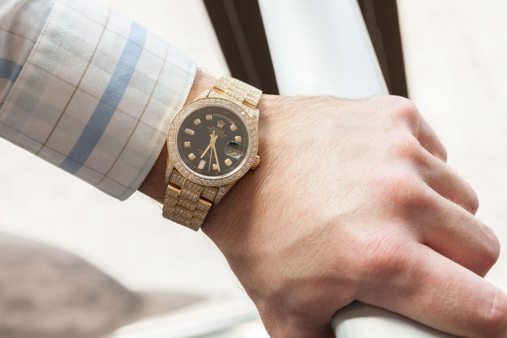 A person's wrist wearing a gold, diamond-studded Rolex Day-Date with a dark face and diamond hour markers, shown against a light plaid shirt.