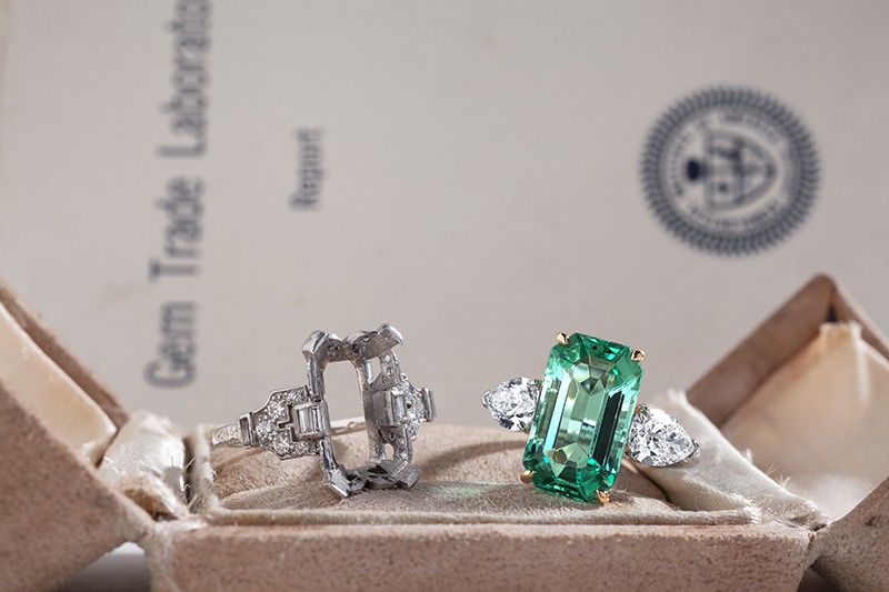 A stunning emerald-cut green gemstone ring flanked by two diamonds, beside a textured jewelry box and a gem lab report.