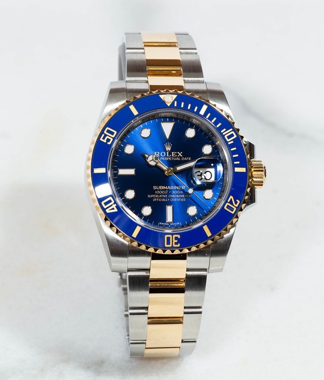 	
A two-tone stainless steel and gold wristwatch with a blue bezel and dial, displaying the time and date, is placed on a white marble surface.
