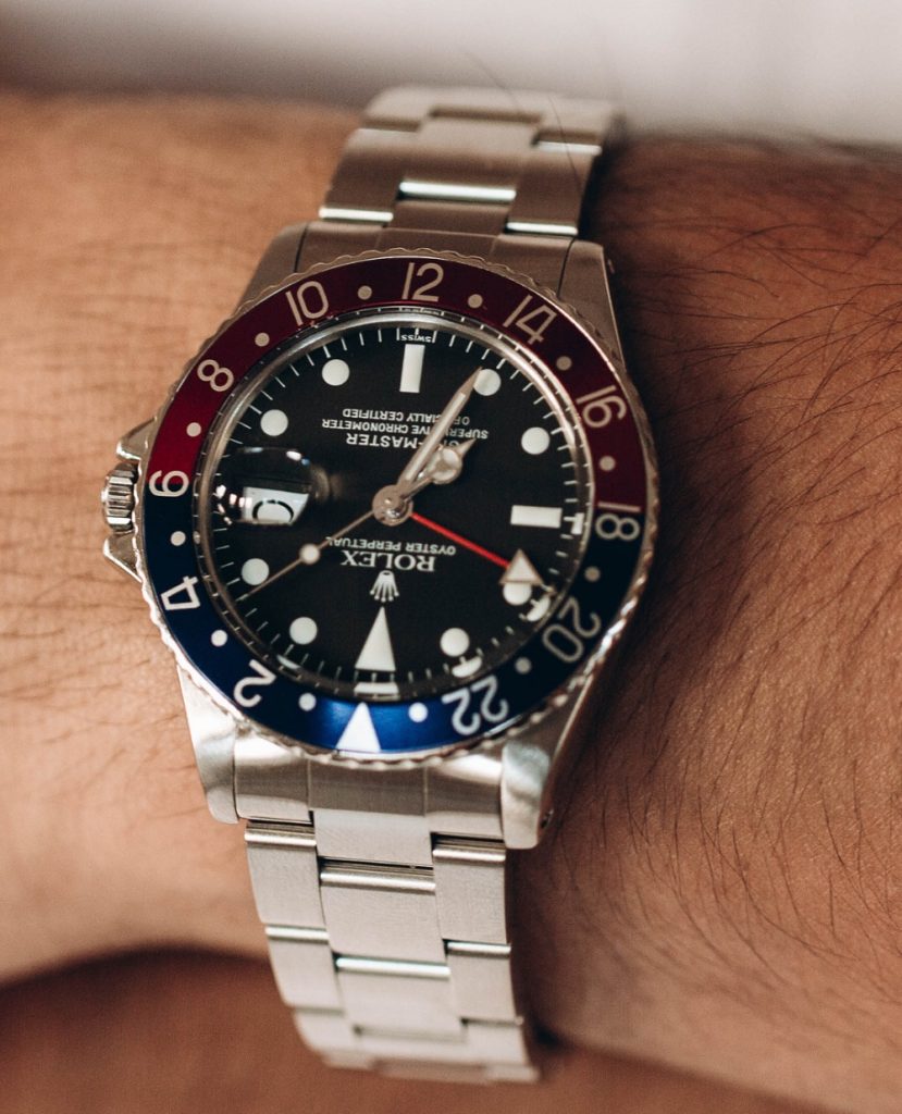 A Rolex Submariner watch with a blue dial and bezel, silver and gold bracelet, is displayed in an open green and wood Rolex box. The iconic luxury timepiece showcases classic Rolex craftsmanship and style.