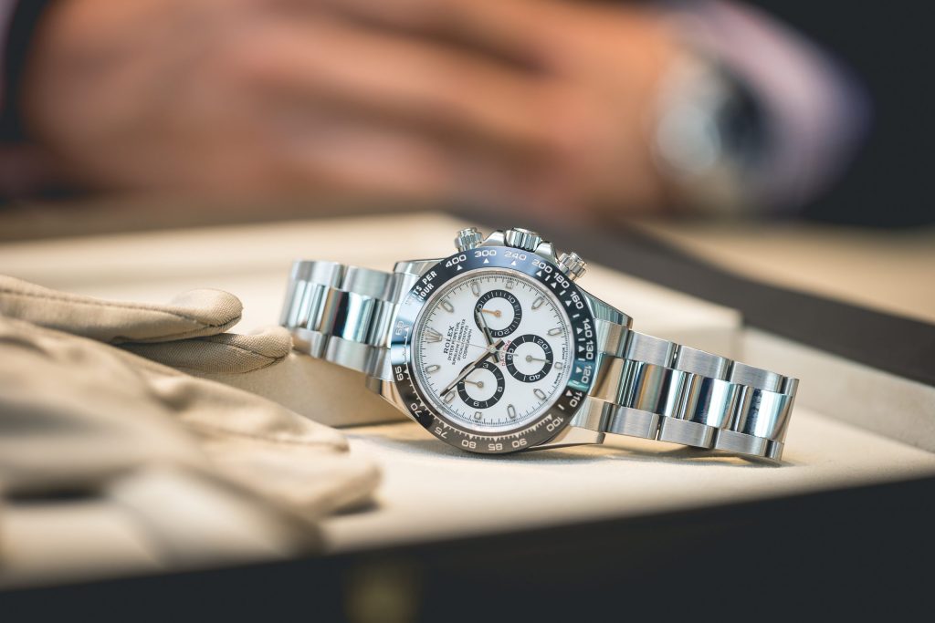 A stainless steel Rolex Daytona watch with a white dial and black subdials is displayed on a tray next to a pair of beige gloves.